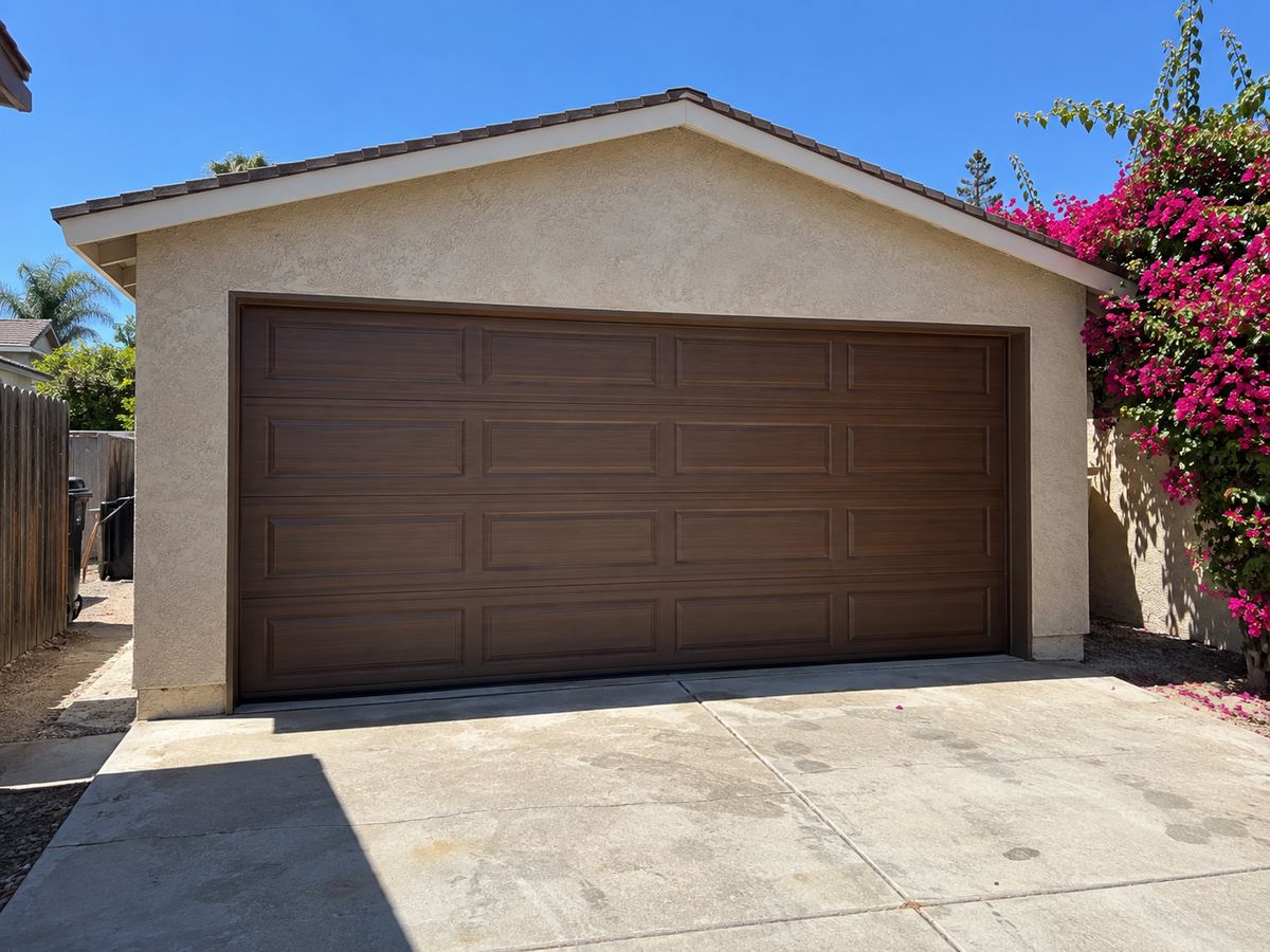 Brown raised-panel double garage door on a detached stucco garage with bougainvillea alongside