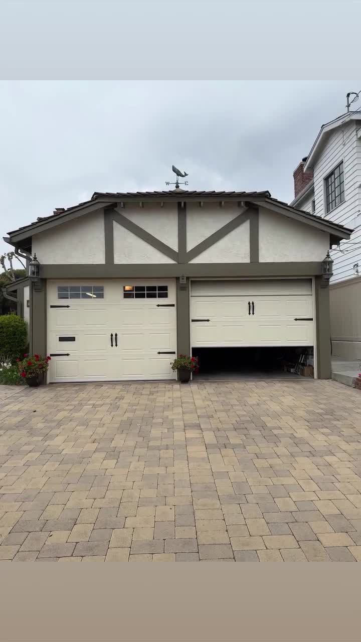 Two cream carriage-house garage doors with dark handles on a Tudor-style home with a paver driveway