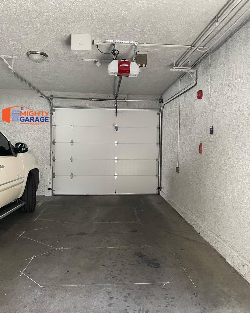 Garage door opener, rail and red emergency release cord mounted on the ceiling above a white single garage door