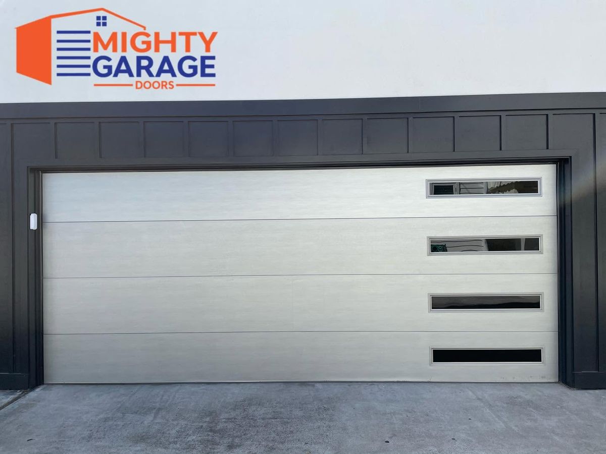 Gray flush-panel modern garage door with a vertical column of narrow windows, framed by dark board-and-batten siding